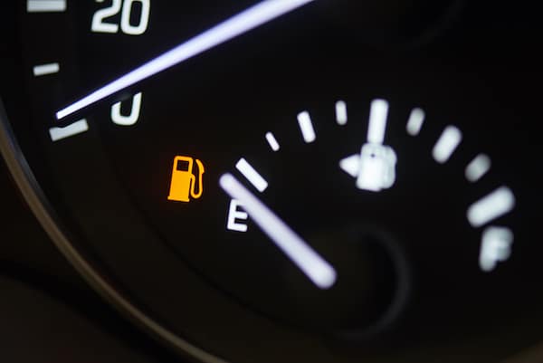 Close-up of a car dashboard fuel gauge showing near empty, with an illuminated yellow low fuel warning light. The gauge needle points towards the "E" (empty) mark.