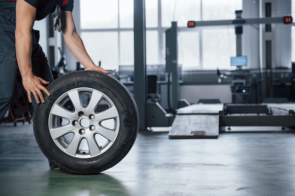 A person with cropped view rolling a car tire inside an automotive workshop. The workshop is equipped with various tools and machinery, and the background shows a vehicle lift and some workstations. The scene has a professional and industrial atmosphere.